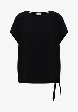 Femme aux cheveux bruns et raides portant un haut noir ample à manches courtes et un jean bleu clair, vue de dos sur fond blanc.