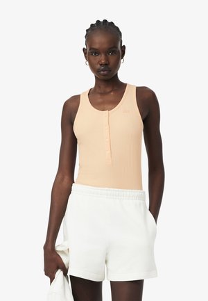 Femme aux cheveux tressés portant un débardeur pêche boutonné et un short blanc, tenant un vêtement blanc dans une main sur un fond uni.