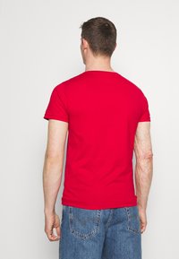 Červené bavlněné tričko s kulatým výstřihem a krátkými rukávy, doplněné modrými džínami. Látka působí hladce, bez viditelných vzorů nebo zdobení.