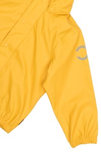 Chaqueta de lluvia amarilla con una superficie suave e impermeable, puños elásticos, cierres a presión y un detalle de logo reflectante en la manga.