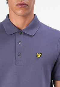 Polo shirt violet en tissu texturé, avec un col, une patte de boutonnage à trois boutons, et un logo d'aigle brodé jaune sur la poitrine.