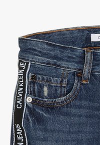 Džínové kalhoty v tmavě modré barvě s poškozenou záplatou. Vybaveny černobílým páskem s logem a oranžovými švy.