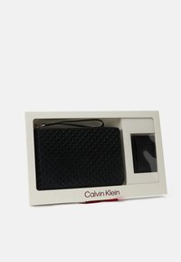 Pochette noire texturée avec motif géométrique et bracelet, présentée dans une boîte blanche avec un porte-cartes noir assorti.