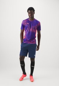 Camisola desportiva da Puma em roxo e rosa, calções azuis marinhos, meias pretas e chuteiras rosas. Apresenta um tecido texturizado e detalhes com o logotipo.