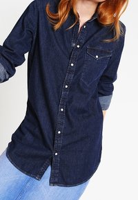 Mörk denimskjorta med knappar, bröstficka, långa ärmar uppvikta vid manschetten och en rundad nederkant. Har en klassisk krage och silverknappar.