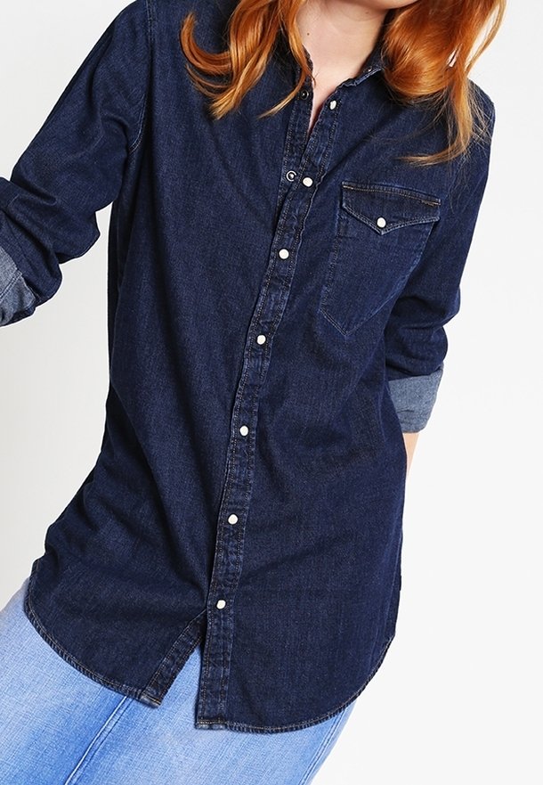 Mörk denimskjorta med knappar, bröstficka, långa ärmar uppvikta vid manschetten och en rundad nederkant. Har en klassisk krage och silverknappar.