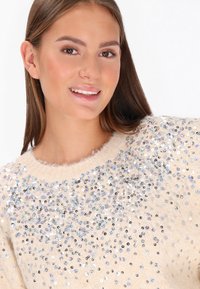 Pulover bej cu o textură moale și pufoasă, decorat cu mărgele multicolore, dispuse prominent pe partea din față. Guler rotund.