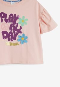 Rosa bomullströja med korta puffärmar, med en lila grafisk text "LEK HELA DAGEN" och färgglada broderade blommor i blått och grönt.