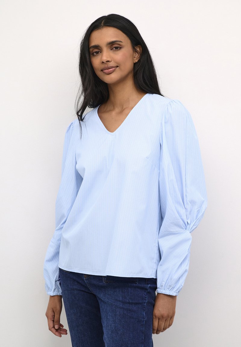 Blusa azzurro chiaro a V con righe verticali, maniche lunghe a sbuffo e polsini arricciati, realizzata in un tessuto morbido, abbinata a jeans blu scuro.