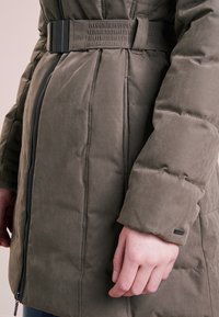 Olivgrön puffjacka med bälte i midjan, långa ärmar och synliga sömdetaljer. Har en framzipper och textilstruktur.