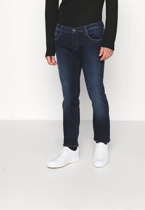 Personne portant un jean slim bleu foncé, un haut à manches longues noir et des baskets blanches, debout sur un sol clair devant un fond uni.