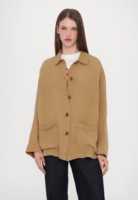 Tan linen oversized shirt with a collar, button-down front, and two front pockets. Long sleeves, relaxed fit, and textured finish.