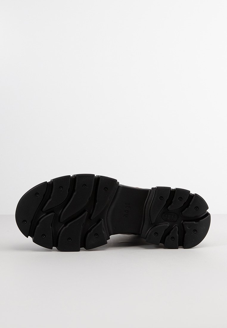 Suela negra y resistente de un zapato con profundas estrías y patrones de agarre circulares, exhibida sobre una superficie y fondo blancos lisos.