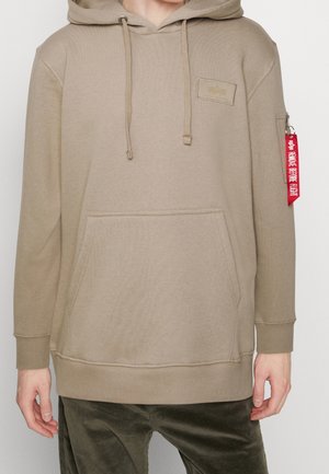 Beige hættetrøje med kængurulomme, ribstrikkede manchetter og et logo-patch. Har en rød mærkat med lynlås fastgjort til ærmet.