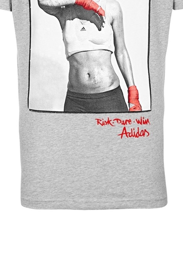 Camiseta de algodón gris con una impresión en blanco y negro de una persona con vendas de boxeo rojas. El texto en la parte inferior dice "Riesgo, Atrevimiento, Victoria" y "Adidas."