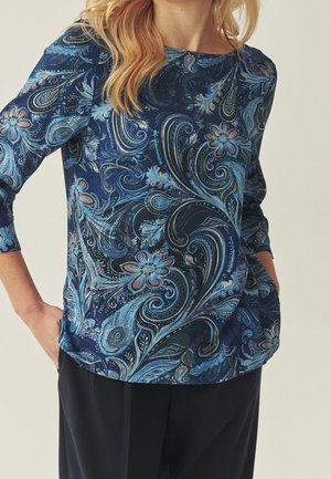 Femme portant une chemise bleu à manches longues avec un motif paisley et floral, les mains dans les poches, associée à un pantalon noir.