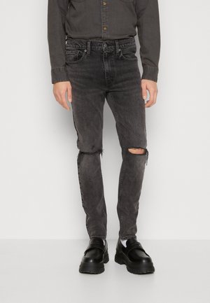 Man draagt vervaagde zwarte gescheurde skinny jeans gecombineerd met zwarte chunky loafers en een donker, dichtgeknoopt overhemd.