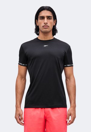 Homme aux cheveux foncés portant un t-shirt noir Reebok et un short rouge vif, debout devant un fond clair uni, face à l'avant.