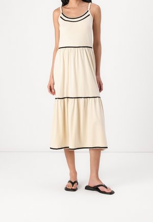 Woman wearing a sleeveless cream midi dress with black trim and black sandals, standing against a plain white background.