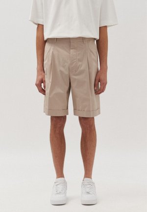 Pantaloni corti beige sartoriali con pieghe, orli risvoltati e tasche laterali. Abbinati a sneakers bianche e una T-shirt bianca dalla vestibilità ampia.