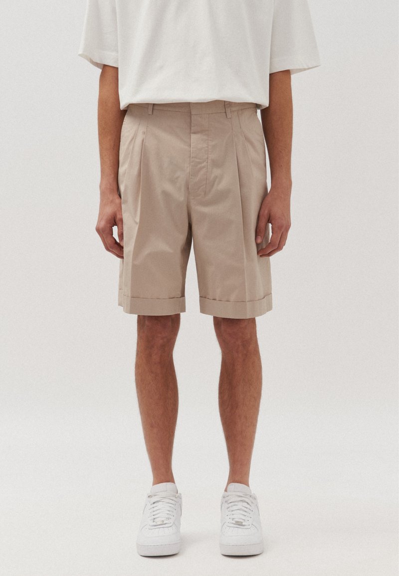 Pantaloni corti beige sartoriali con pieghe, orli risvoltati e tasche laterali. Abbinati a sneakers bianche e una T-shirt bianca dalla vestibilità ampia.