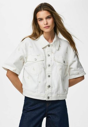 Jeune femme aux longs cheveux portant une veste blanche à manches courtes boutonnée et un jean bleu foncé sur un fond uni.