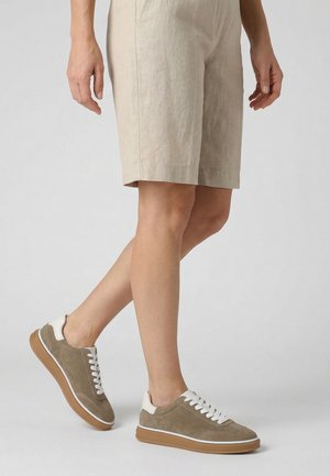 Person iført beige knælange shorts og lysebrune ruskindssneakers med hvide snørebånd og såler, stående mod en ensfarvet baggrund.