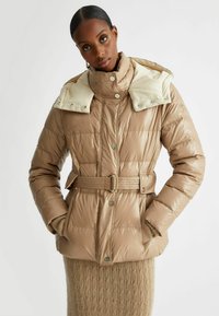 Giacca a vento beige con vita cintata, colletto alto e cappuccio crema a contrasto. Tessuto testurizzato con motivi trapuntati e bottoni metallici.