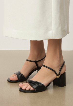 Fekete bőr magassarkú szandál széles, csavart pánttal a lábujjak felett, vékony bokapántokkal és blokk sarokkal, amely arany díszítést tartalmaz.