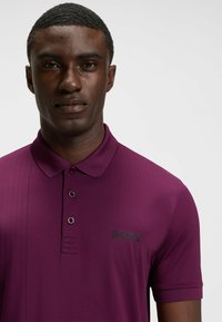 Lila Poloshirt mit Kragen, fünfknöpfiger Leiste, dezenten vertikalen Streifen auf der linken Seite und schwarzem BOSS-Logo auf der Brust.