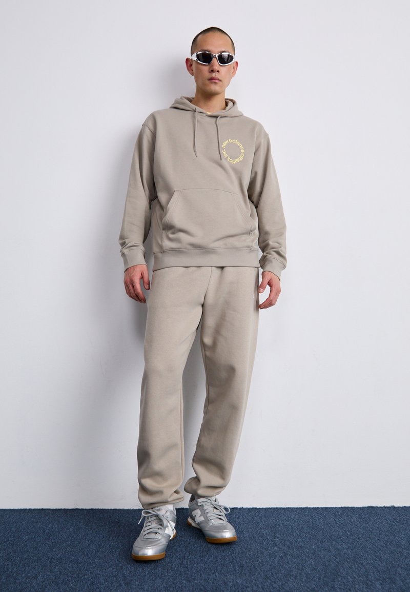Beiger Hoodie mit einer Vordertasche und Logo, kombiniert mit passenden beigen Jogginghosen. Silberne Sportschuhe mit grauen Schnürsenkeln und Gummisohle.