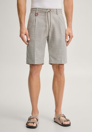 Mann trägt hellgraue Shorts mit Kordelzug, Falten und Metallenden, kombiniert mit beigen Sandalen mit offenen Zehen und schwarzen Schnallen.