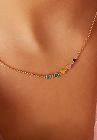 Goldene Halskette mit abwechselnden herzförmigen Anhängern in Türkis, Grün, Gelb, Pink und Lila, vor einem hautfarbenen Hintergrund.