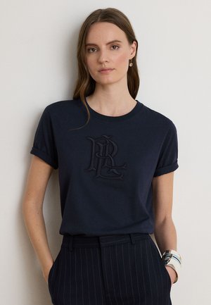 Kvinde med langt brunt hår iført en marineblå T-shirt med broderet logo, bukser med nålestriber, guldøreringe og et hvidt mønstret armbånd.