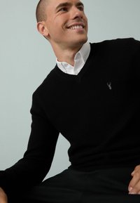 Schwarzer, strukturierter V-Ausschnitt-Pullover aus Rippmaterial, getragen über einem weißen Hemd mit Kragen, mit einem kleinen grauen gestickten Logo auf der Brust.