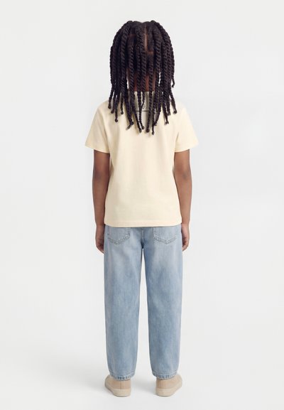 Παιδί με μακριά πλεξούδα στέκεται με την πλάτη, φορώντας μπλούζα με κοντά μανίκια σε κρεμ απόχρωση, ανοιχτό μπλε τζιν και μπεζ παπούτσια, σε απλό λευκό φόντο.