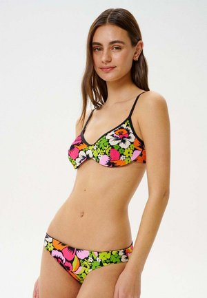 Młoda kobieta w kolorowym kwiecistym bikini z zielonymi, różowymi, pomarańczowymi i białymi wzorami, delikatnie uśmiechająca się na jednolitym białym tle.