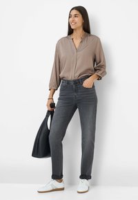 Grijze hoge taille jeans, relaxte pasvorm, gecombineerd met een taupe blouse en witte sneakers. Zwarte totebag in één hand.