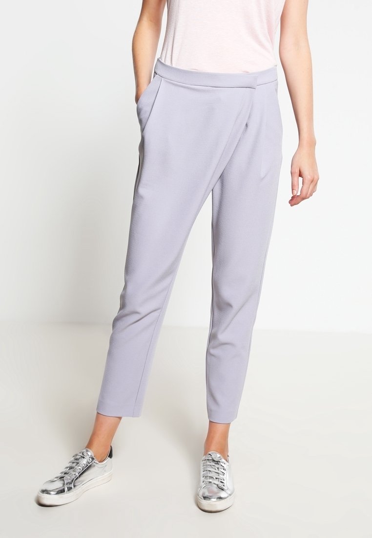 Femme portant un pantalon fuselé lilas clair avec poches latérales et des baskets métalliques argentées, se tenant devant un fond blanc uni.