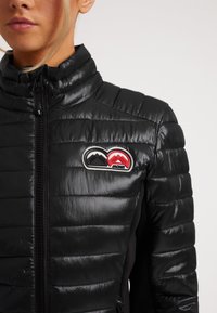Giacca nera imbottita con colletto alto, chiusura con zip e patch logo rosso e nero sul petto. Il materiale è lucido e testurizzato.
