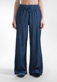 Osoba w luźnych, ciemnoniebieskich spodniach o szerokich nogawkach z elastycznym pasem i sznurkiem, stojąca z rękami swobodnie opuszczonymi wzdłuż ciała.