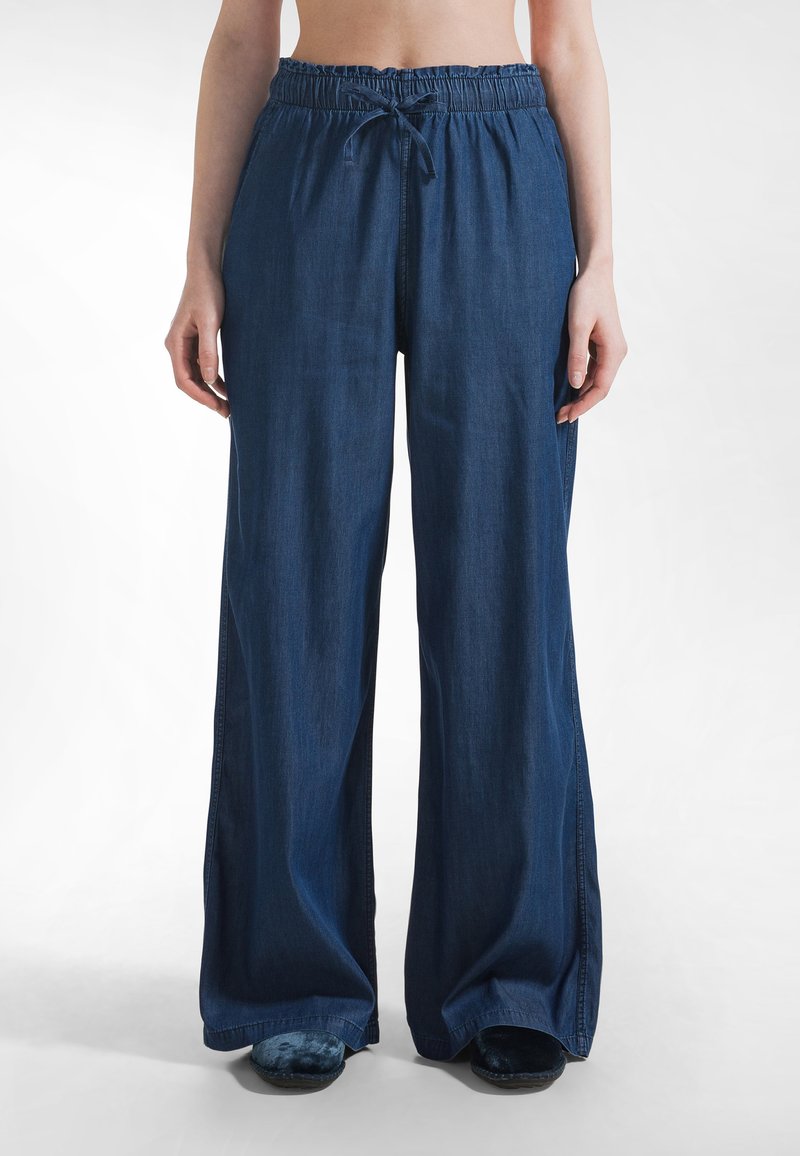 Osoba w luźnych, ciemnoniebieskich spodniach o szerokich nogawkach z elastycznym pasem i sznurkiem, stojąca z rękami swobodnie opuszczonymi wzdłuż ciała.