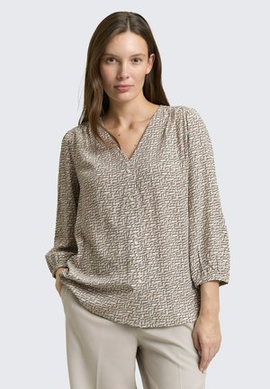 Femme portant un chemisier beige à motifs avec col en V et manches 3/4 froncées, ainsi qu'un pantalon beige clair, debout avec une main dans la poche.