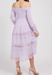 Lila schulterfreies Kleid mit gerafftem Oberteil, langen durchsichtigen Ärmeln, gestuften Rüschen und Spitze am Saum. Getragen mit roségoldenen Absätzen.