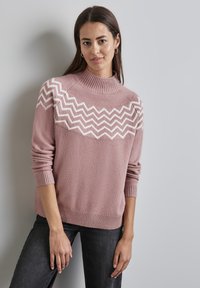 Rosa Pullover mit hohem Kragen, der ein weißes Zickzackmuster über die Brust aufweist. Weiche Textur, gerippte Bündchen und Saum. Genossen mit dunklen Jeans.