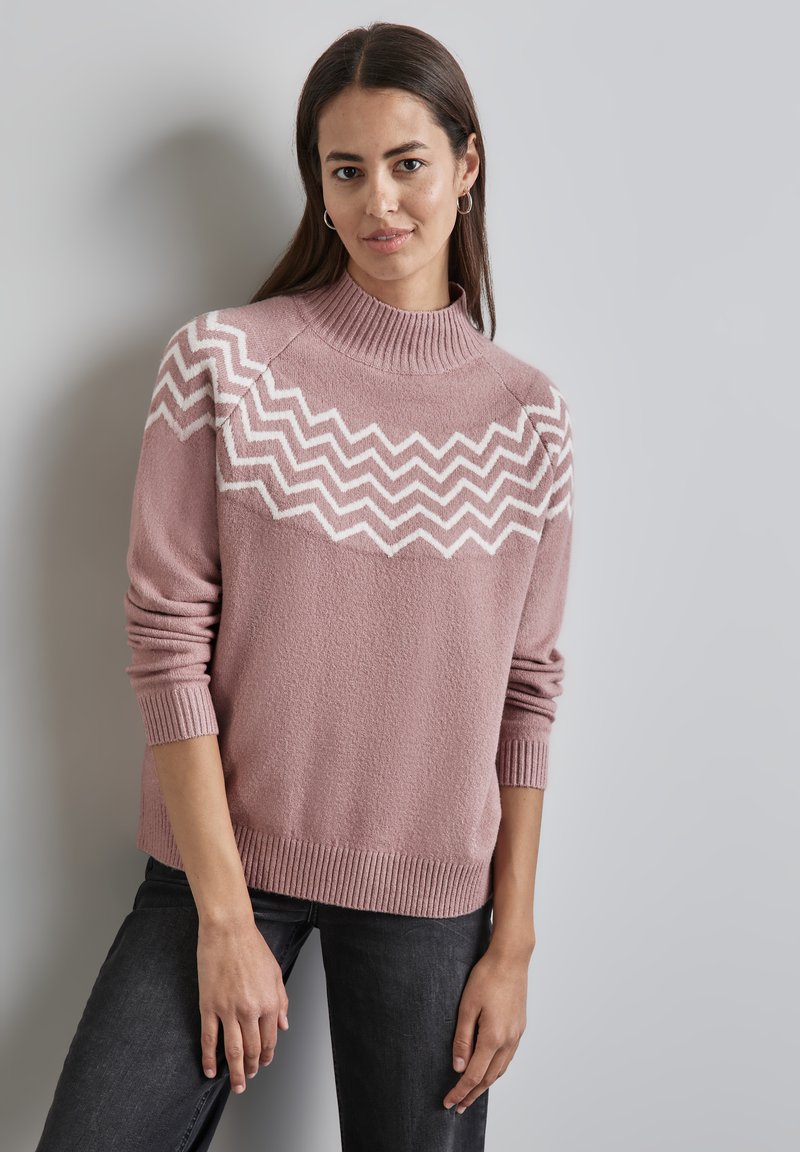 Rosa Pullover mit hohem Kragen, der ein weißes Zickzackmuster über die Brust aufweist. Weiche Textur, gerippte Bündchen und Saum. Genossen mit dunklen Jeans.