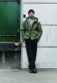 Groen geïsoleerd jack met twee zakken, hoge kraag, gedragen over een grijze trui met textuur, gecombineerd met een zwarte broek en zwarte schoenen.