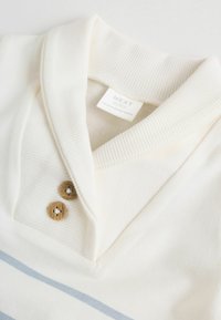 Bela pletena polo majica z rebrastim ovratnikom in dvema lesenima gumboma. Na telesu so svetlo modre vodoravne črte. Oznaka je vidna na notranji strani.