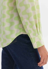 Green and cream blouse with wavy pattern, long sleeves, buttoned cuffs, and a curved hem, paired with dark denim jeans.