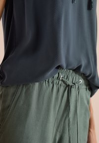 Personne portant un haut sans manches foncé rentré dans un pantalon vert ample à cordon, taille et partie supérieure des hanches visibles.